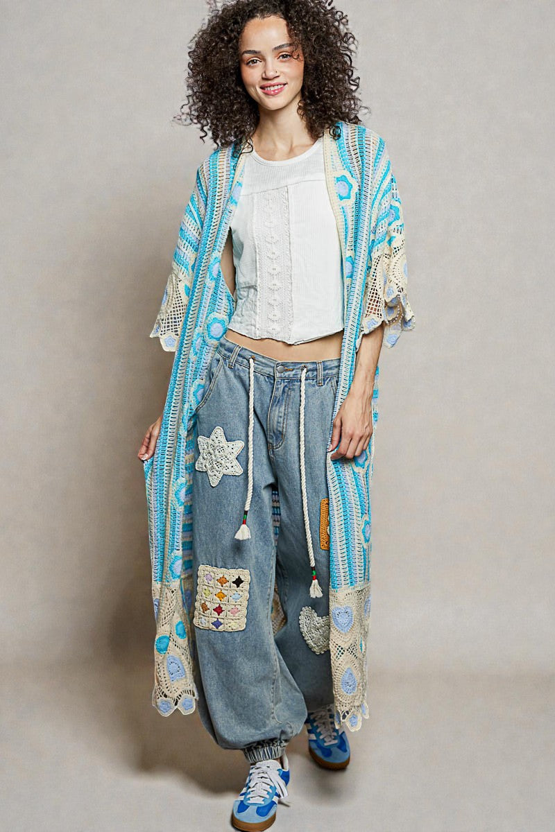 Woman wearing a colorful kimono-style jacket with floral patterns and denim pants on a plain background