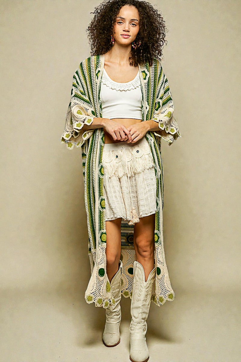 Woman wearing a green and white patterned cardigan over a white dress with lace details, standing against a beige background.