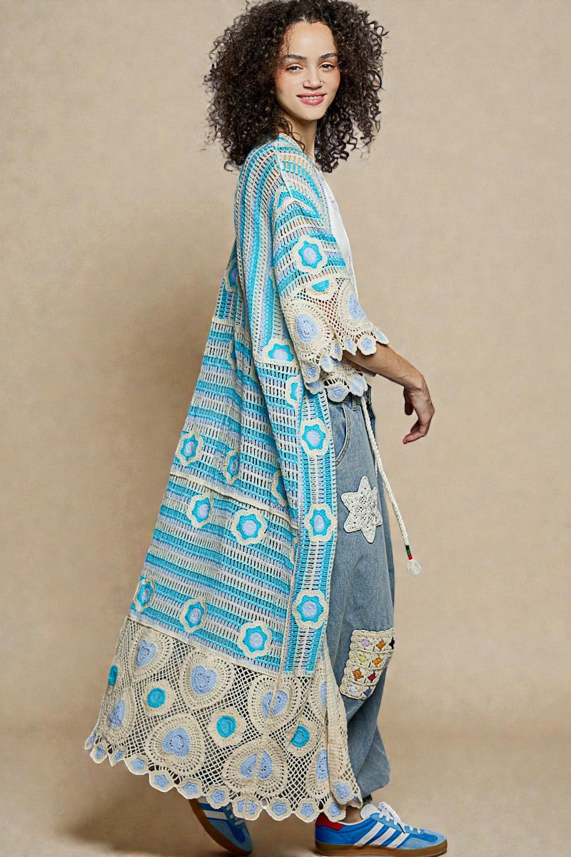 Woman wearing a long, patterned blue and white garment with denim pants on a beige background