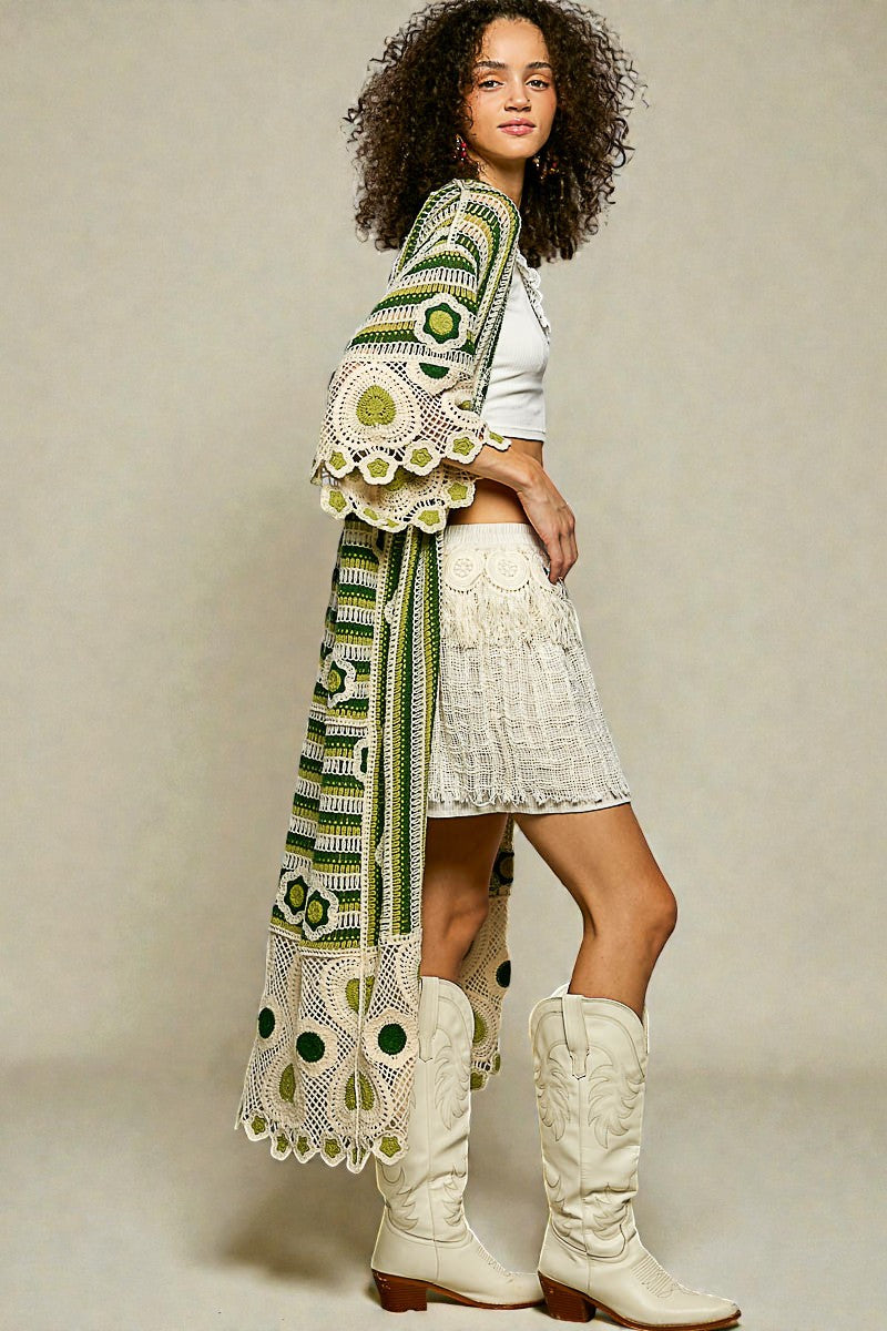 Woman wearing a long, patterned green and white shawl over a white outfit with white boots on a neutral background