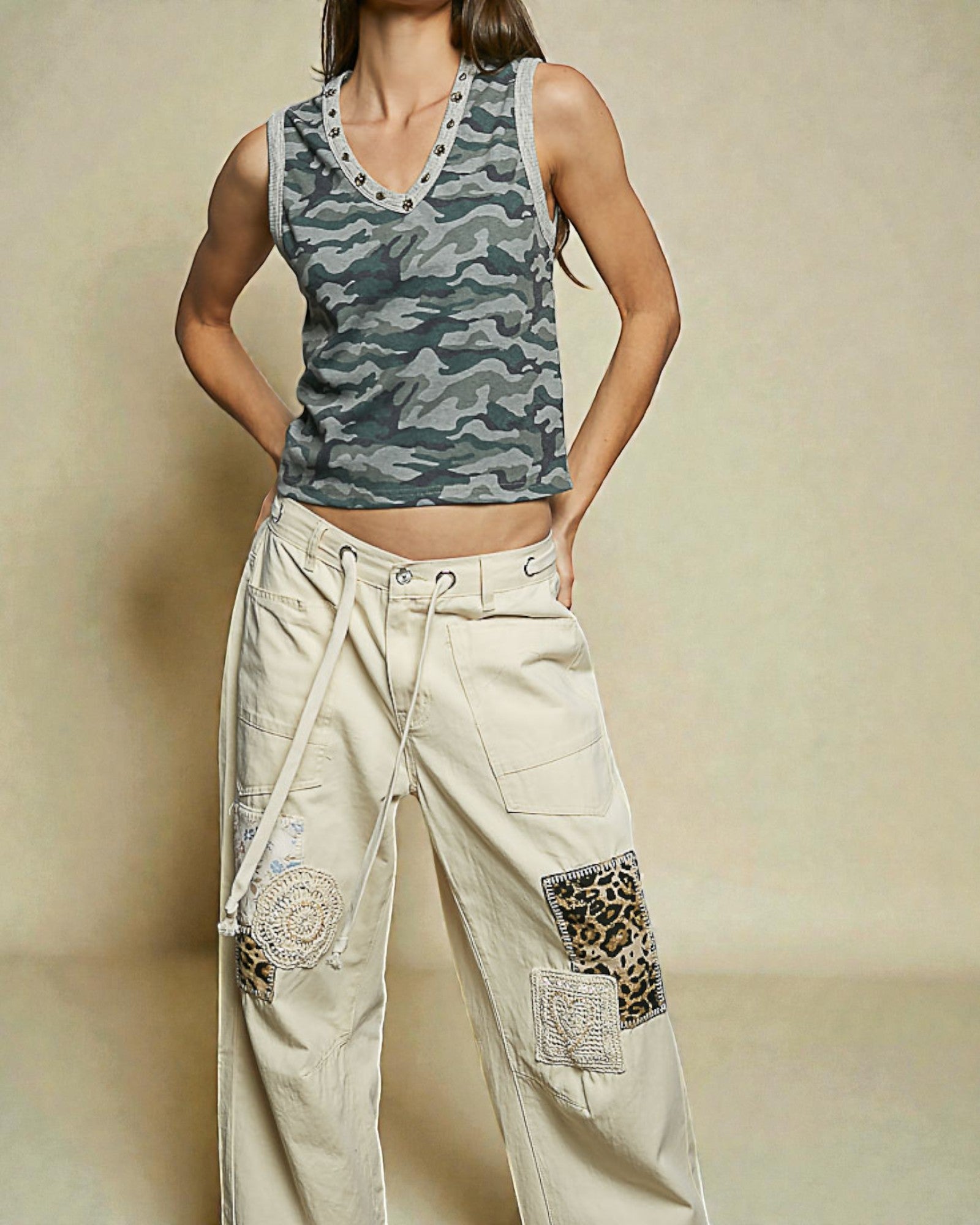Person wearing a camouflage tank top and beige pants with patches on a beige background