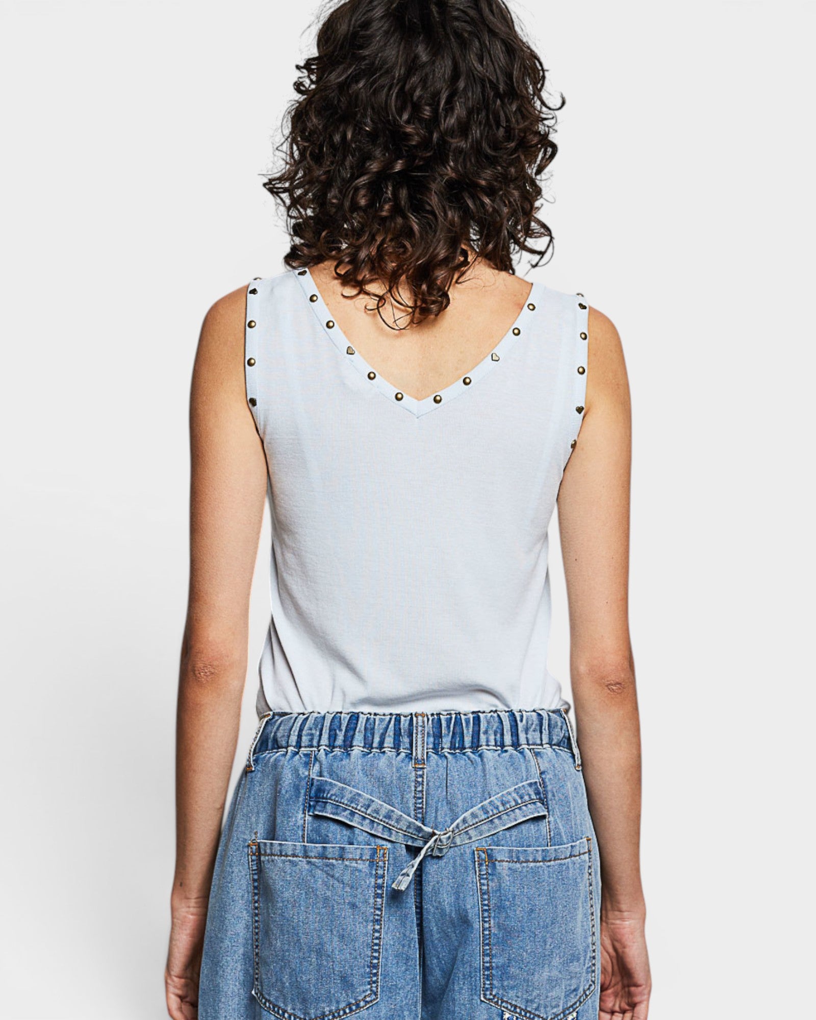 Person wearing a light blue sleeveless top with studded details and blue jeans on a white background