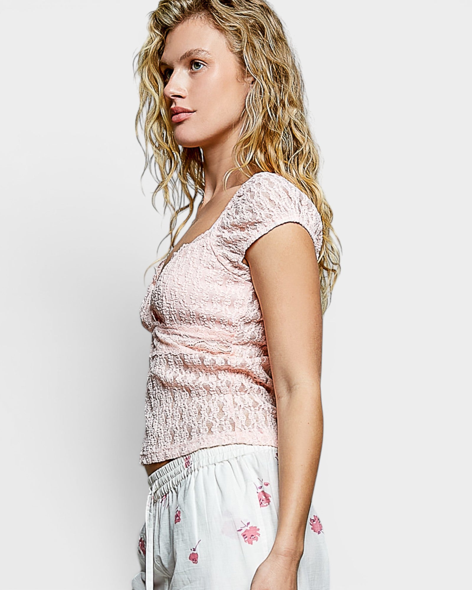 Woman wearing a pink lace top and white pants with floral embroidery on a light gray background