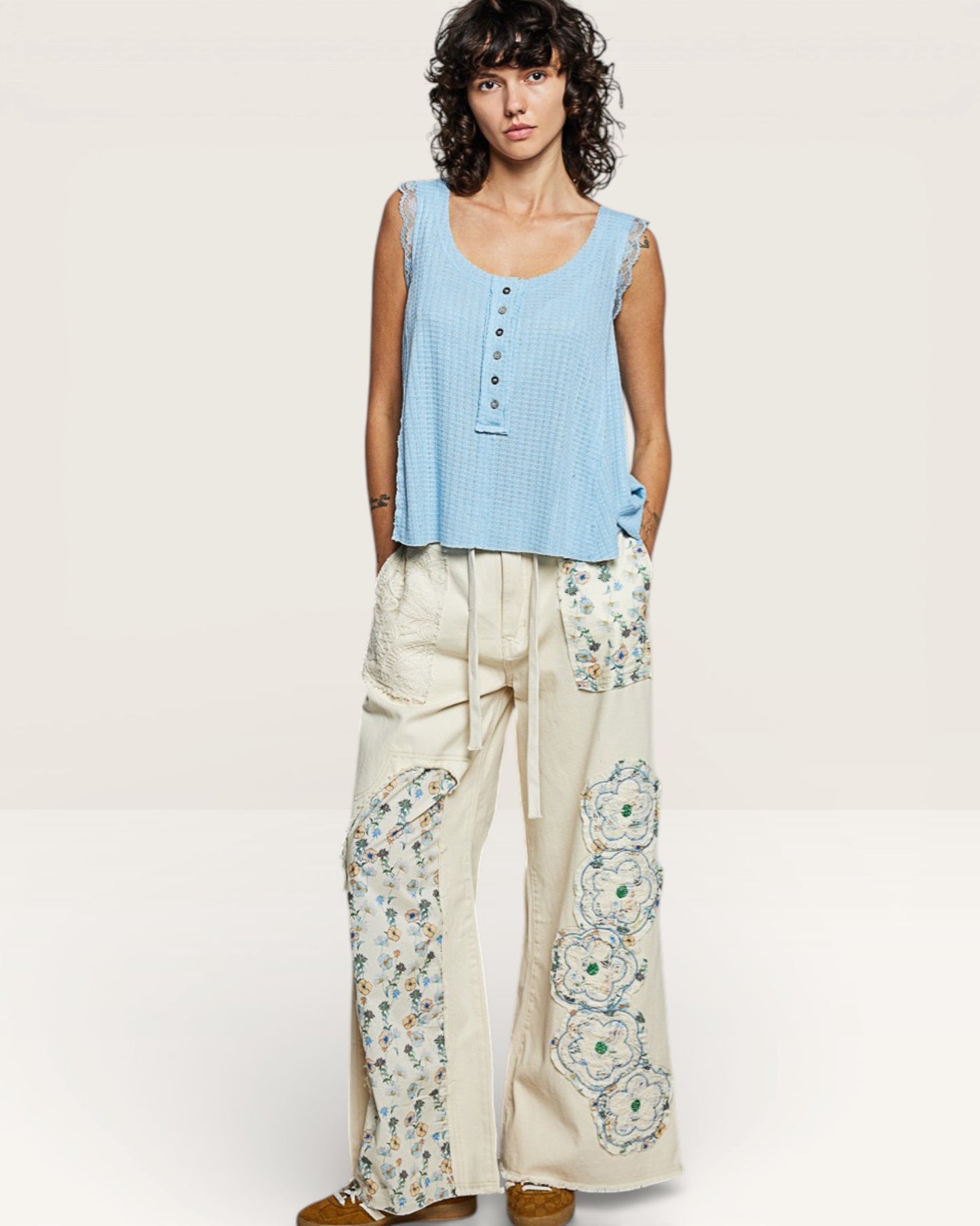 Woman wearing a light blue sleeveless top and cream pants with floral patterns on a white background