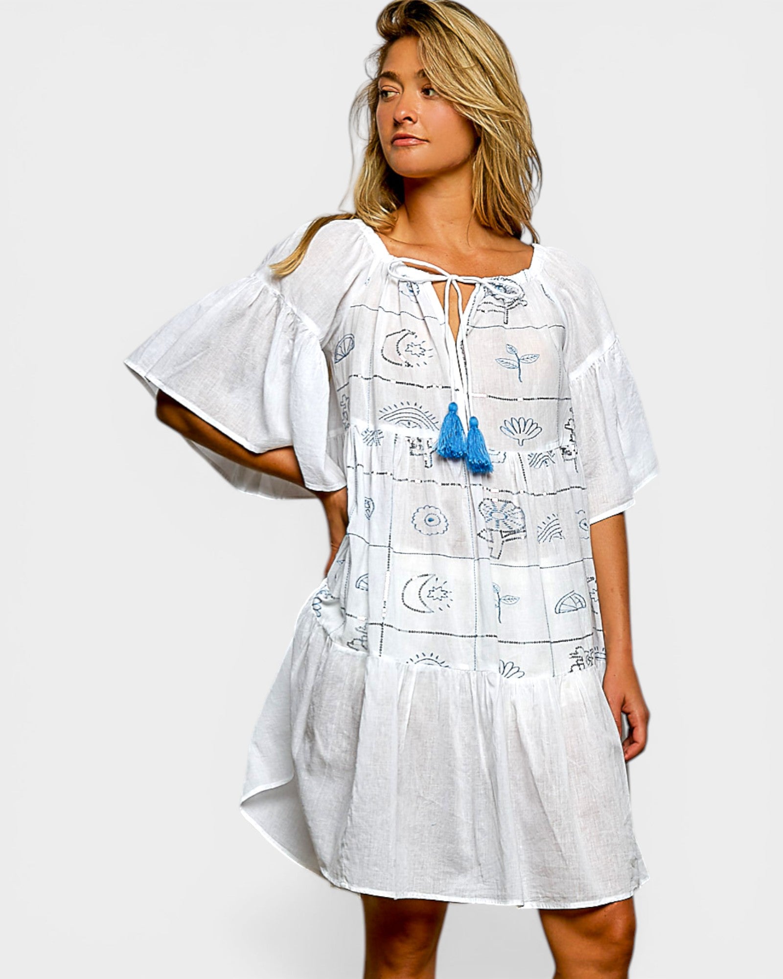 Woman wearing a white dress with blue patterns and tassels on a plain background