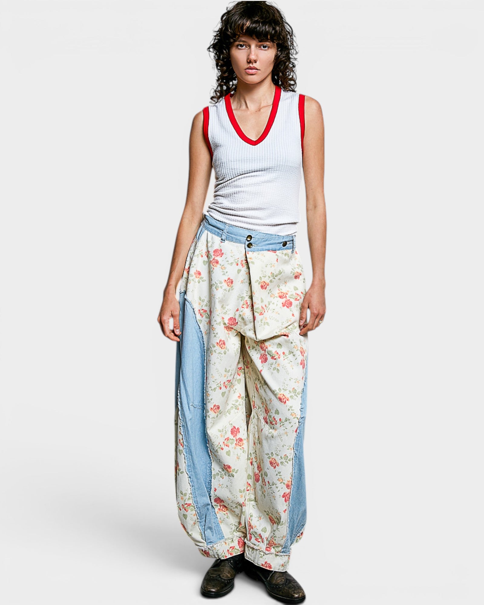 Woman wearing a white tank top with red trim and floral harem pants on a plain background