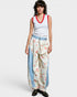 Woman wearing a white tank top with red trim and floral harem pants on a plain background