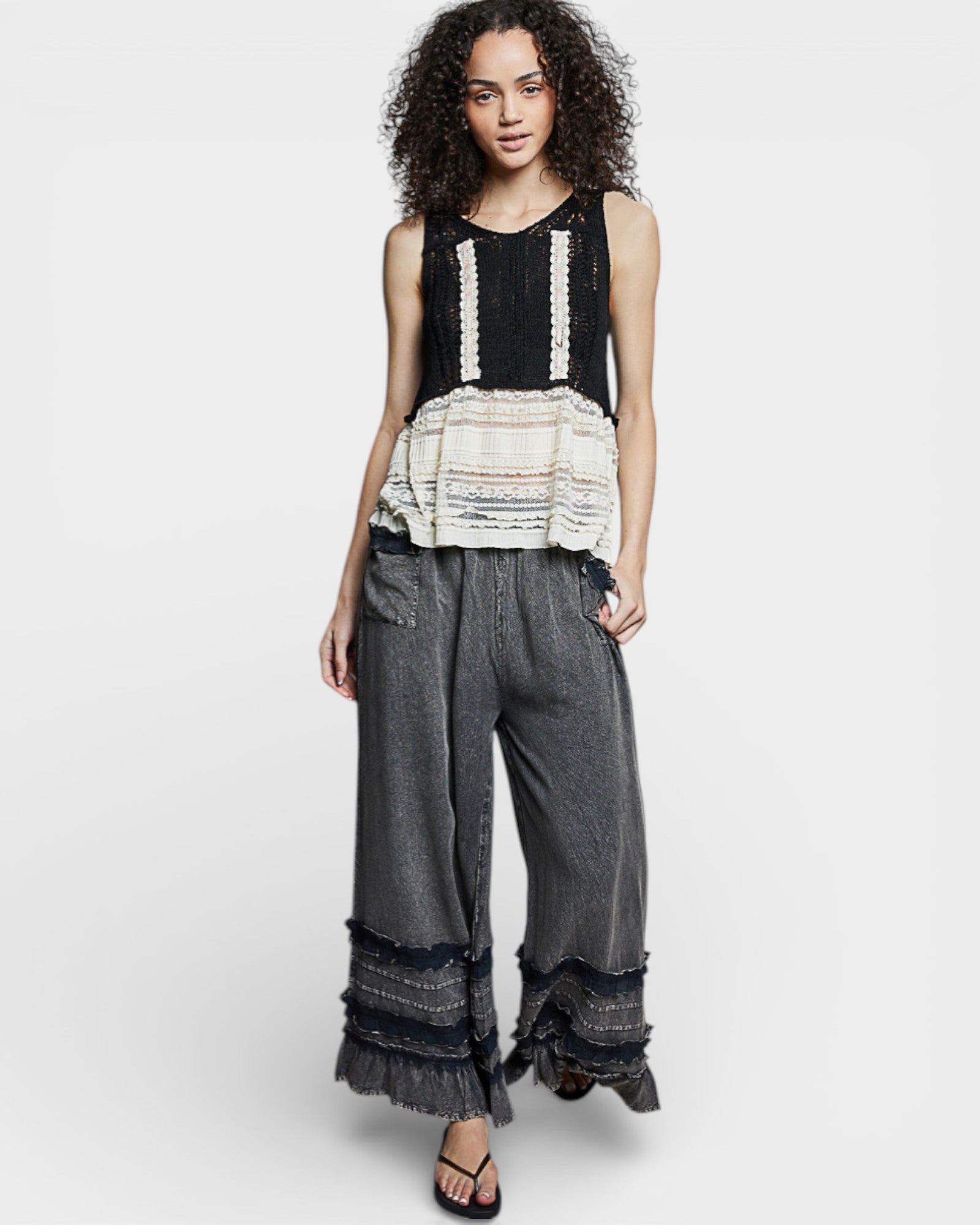 Woman wearing a black sleeveless top with ruffled details and gray wide-leg pants on a white background