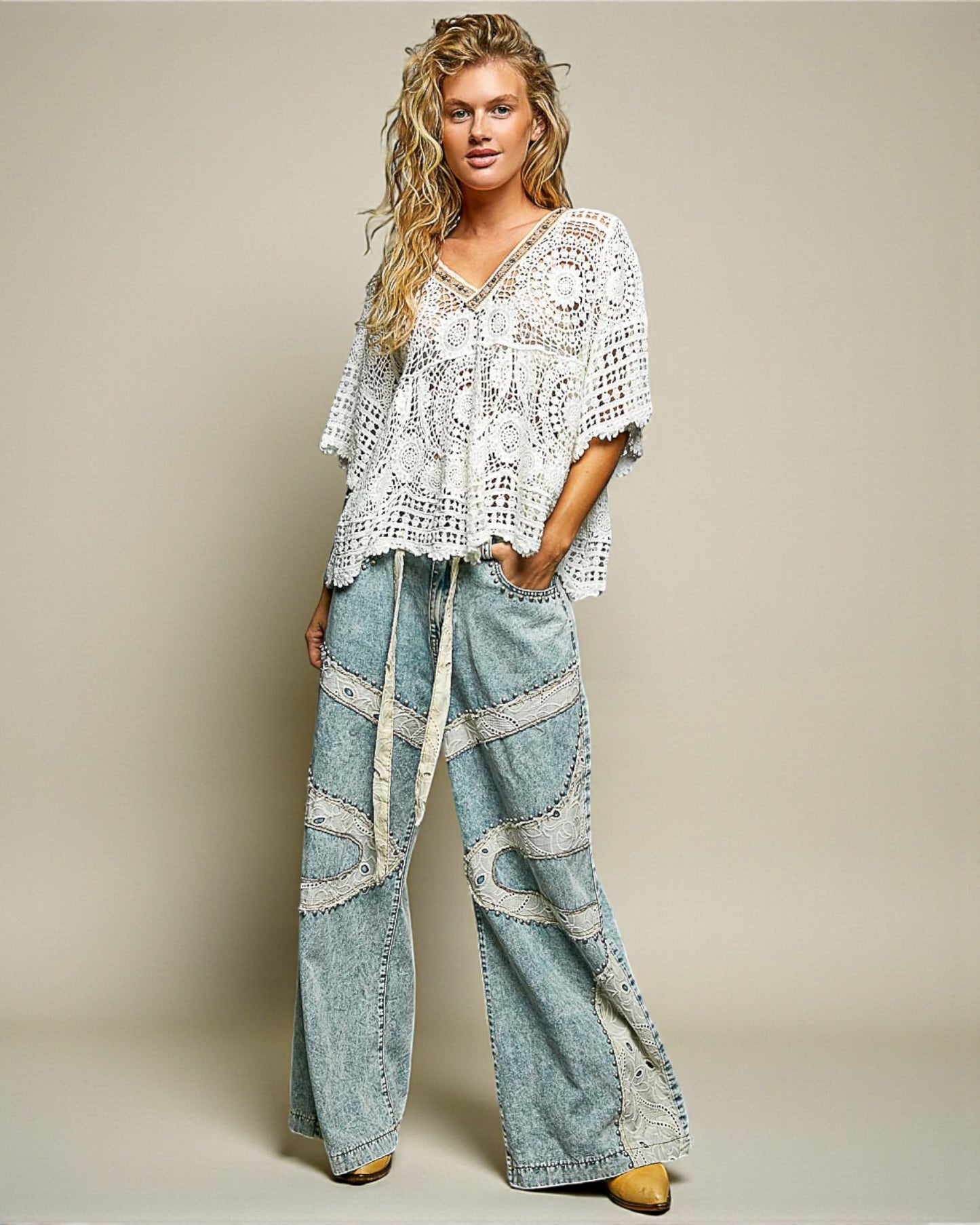 Woman wearing a white lace top and light blue denim pants against a beige background