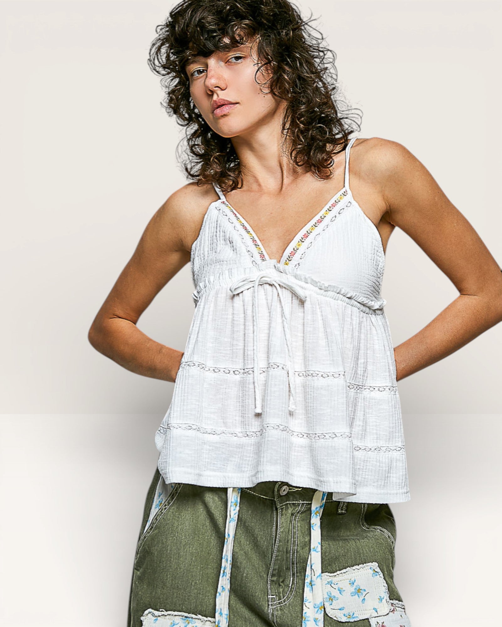 Woman wearing a white lace top and green pants with a floral apron against a plain background