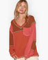 A woman wearing a long-sleeve knit sweater with a color-blocked design in rust orange, terracotta brown, and coral pink, featuring a chest pocket and contrast stitching.