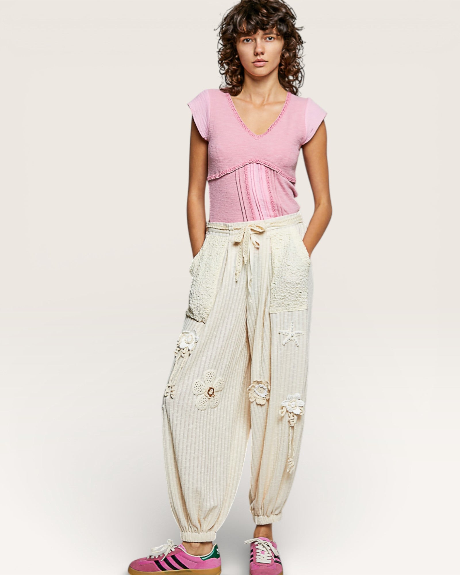 Woman wearing a pink top and white pants with floral details on a plain background