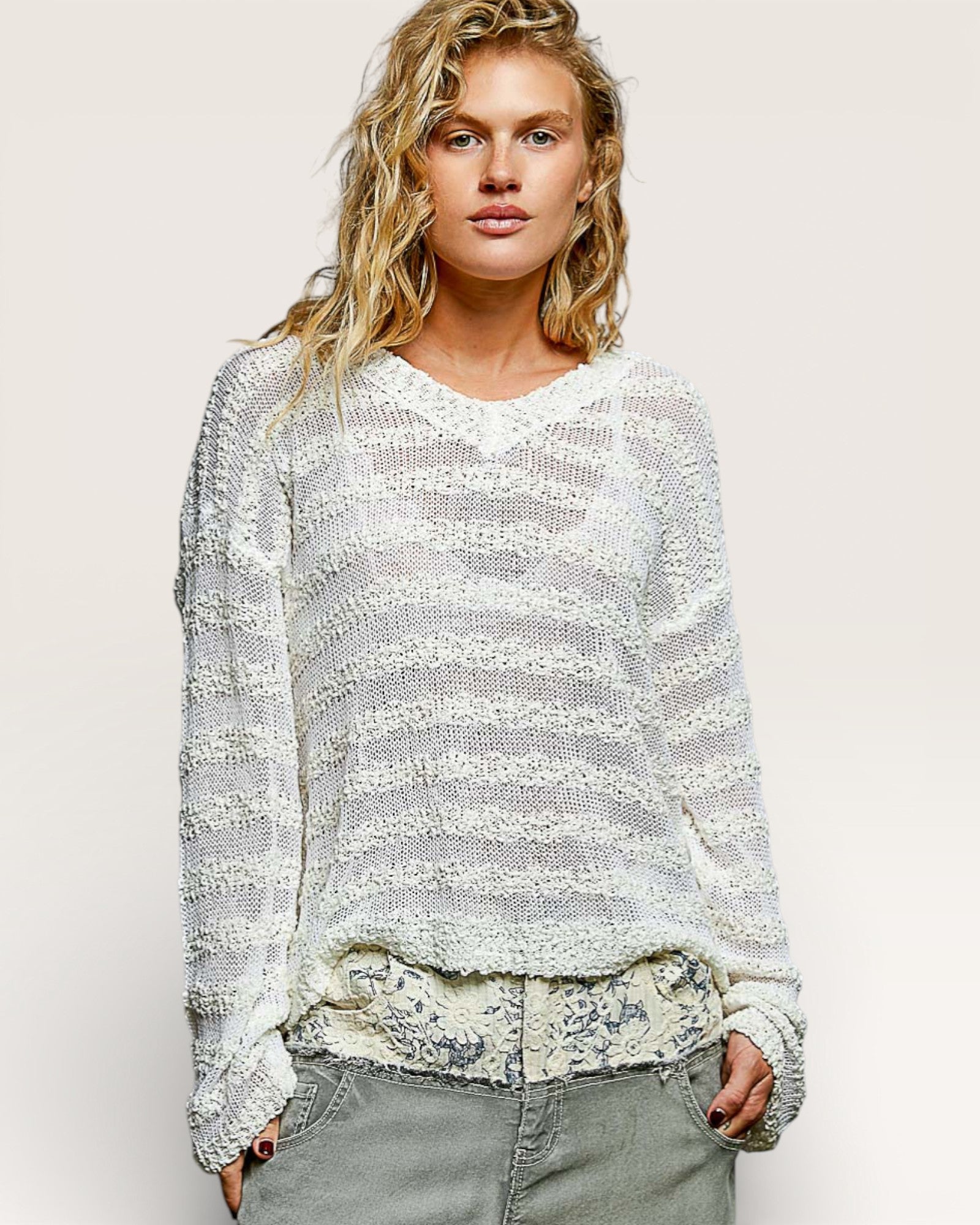Woman wearing a textured white sweater over a floral top with gray pants on a plain background