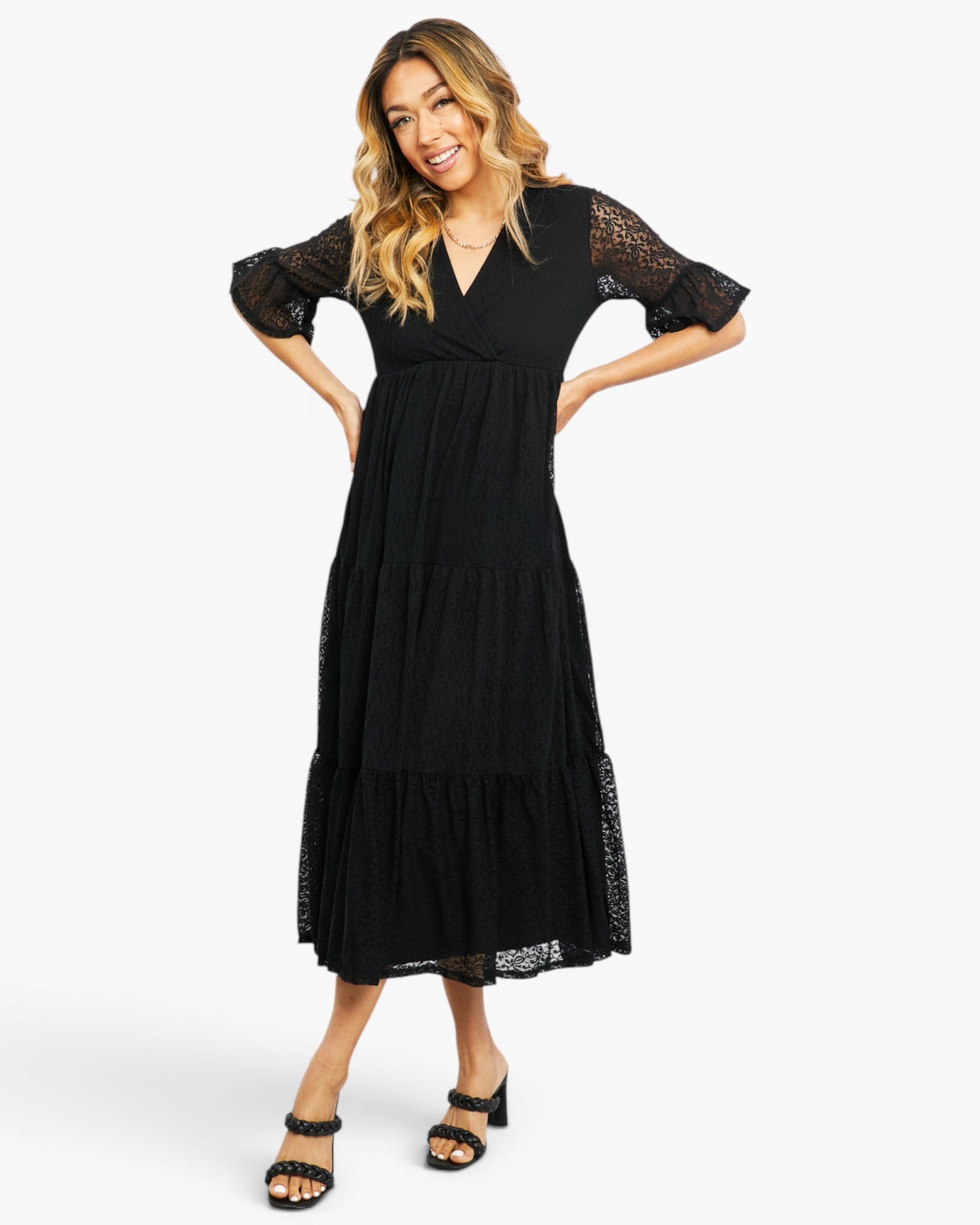 A woman modeling a black midi dress with sheer lace flutter sleeves and a tiered skirt, paired with black heeled sandals, against a plain white studio background.