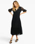 A woman modeling a black midi dress with sheer lace flutter sleeves and a tiered skirt, paired with black heeled sandals, against a plain white studio background.
