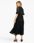 A woman modeling a black midi dress with sheer lace flutter sleeves and a tiered skirt, shown from the back against a light gray studio background.