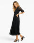 A woman modeling a black midi dress with sheer lace short sleeves and side pockets, paired with black strappy heels, against a plain white studio background.