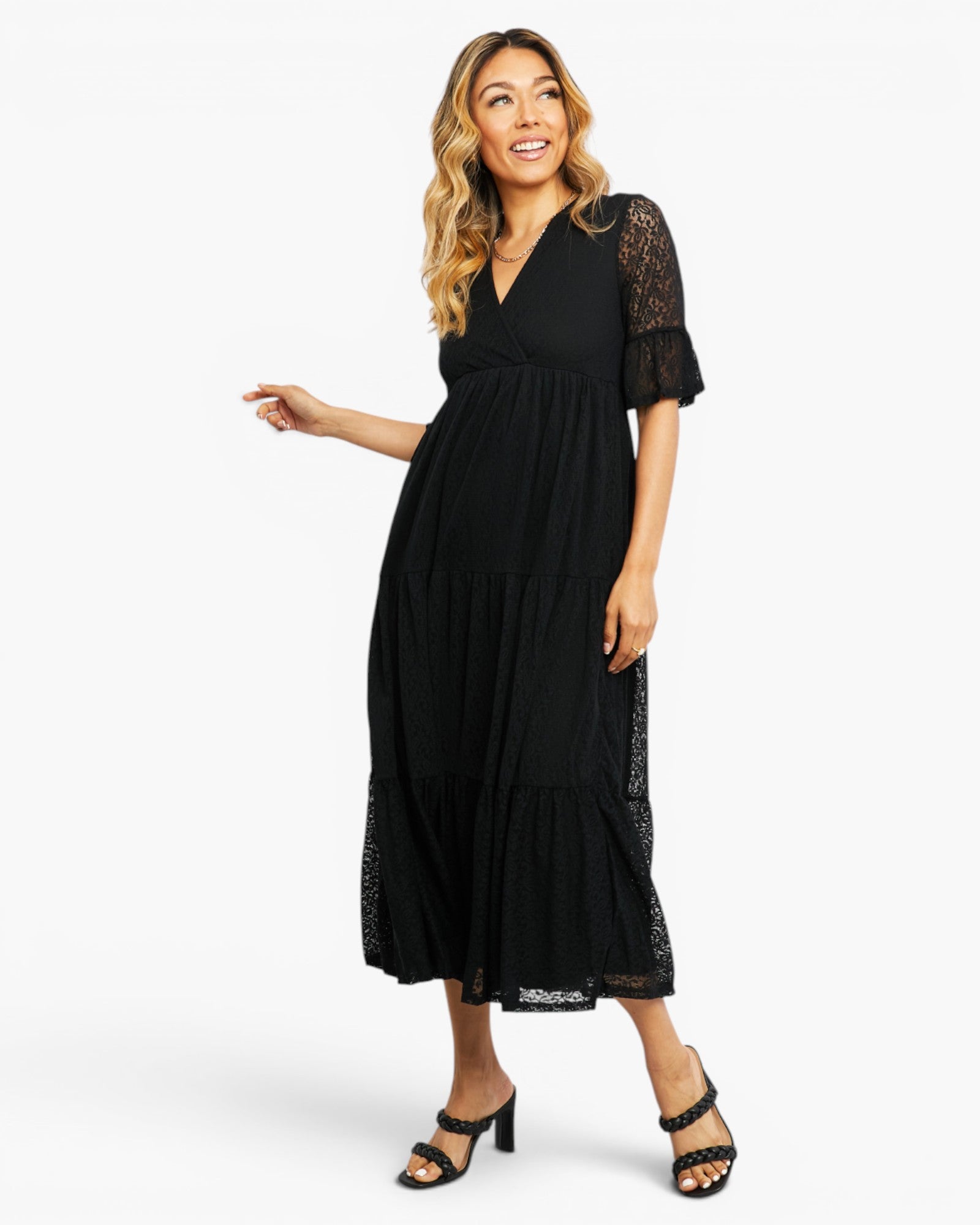 A woman modeling a black midi dress with sheer lace flutter sleeves and a tiered skirt, paired with black heeled sandals, against a plain white studio background.