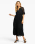 A woman modeling a black midi dress with sheer lace flutter sleeves and a tiered skirt, paired with black heeled sandals, against a plain white studio background.