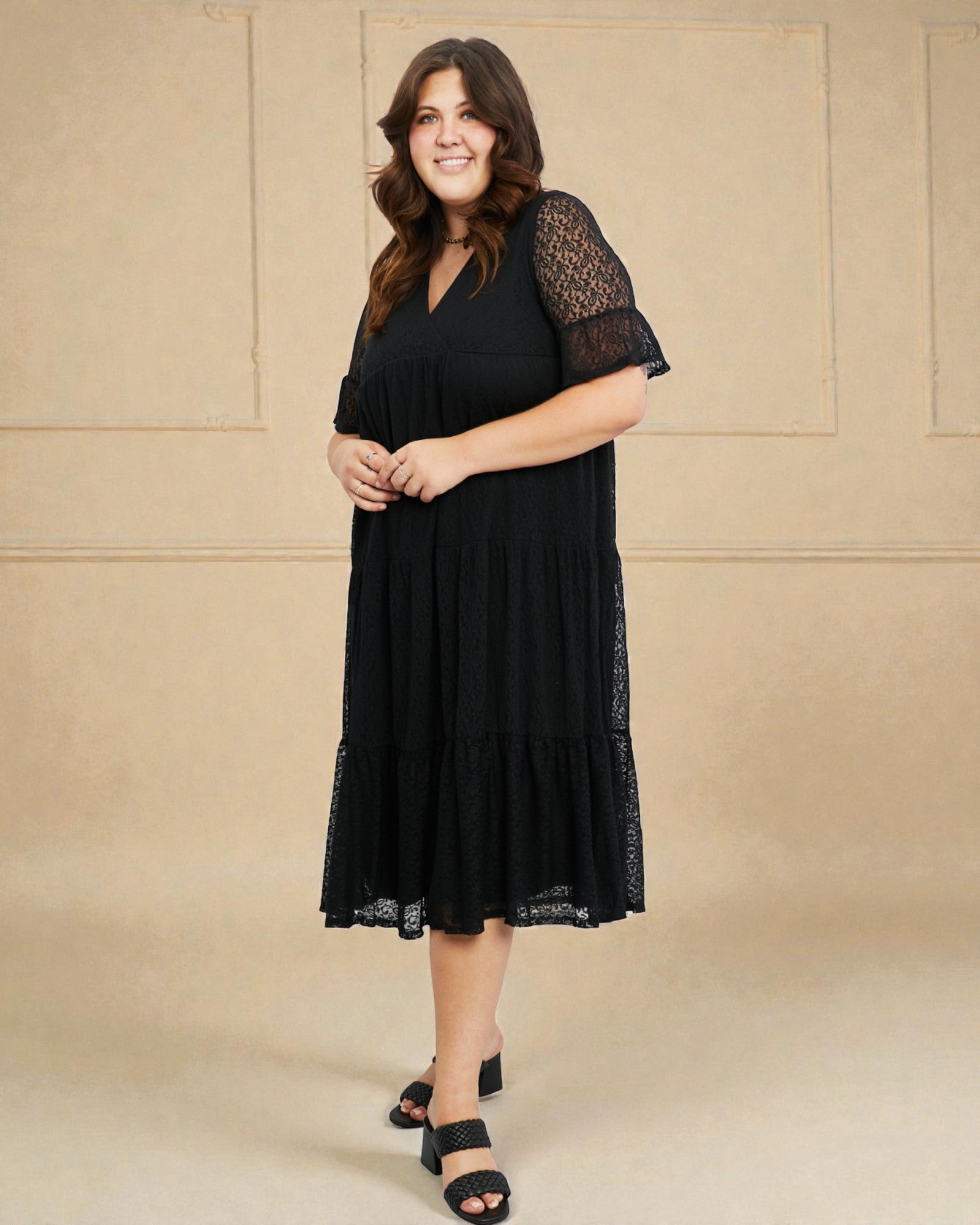 Woman wearing a black dress with lace sleeves against a beige wall.