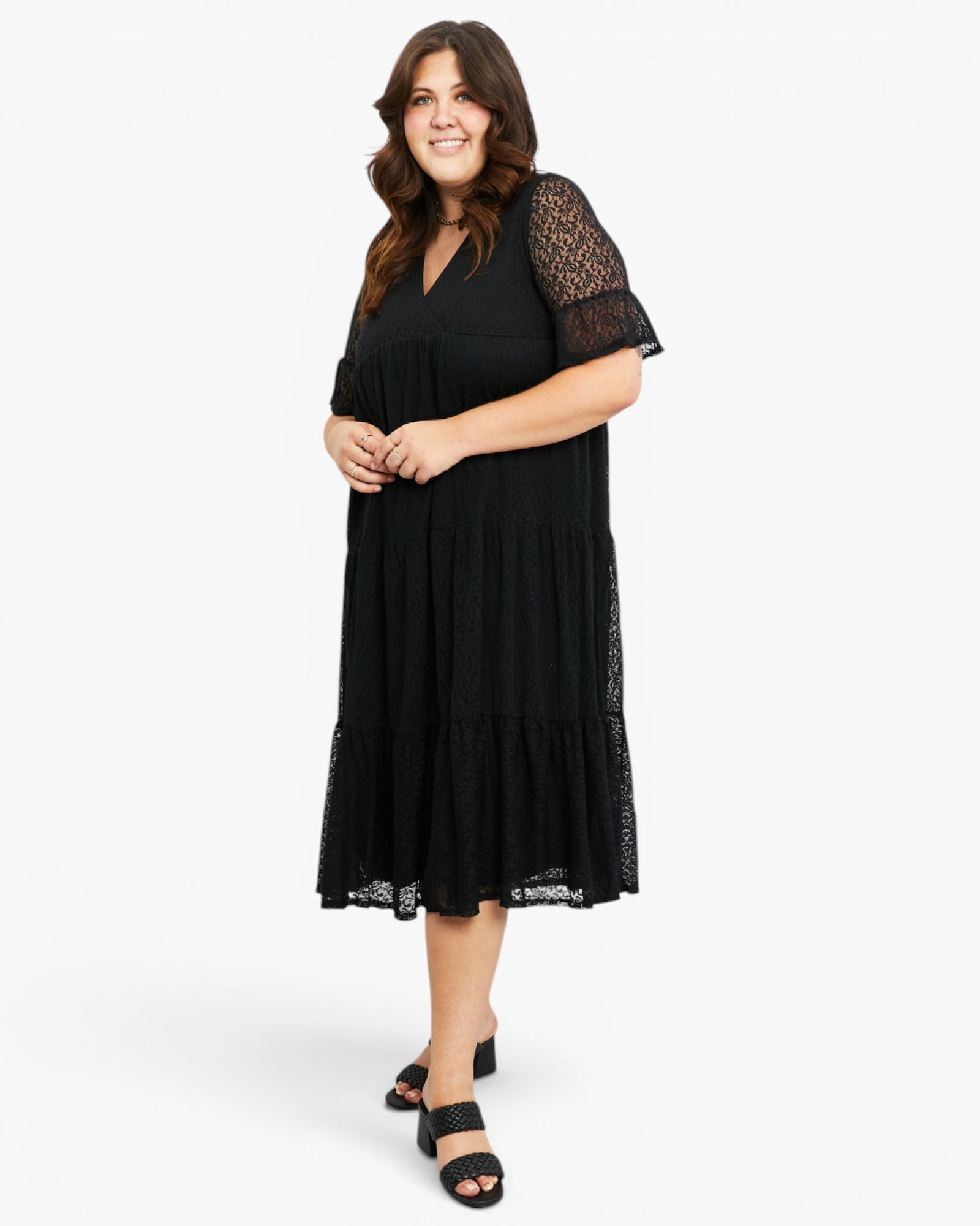 A woman modeling a black midi dress with sheer lace flutter sleeves and a tiered skirt, paired with black slide sandals, against a plain white studio background.
