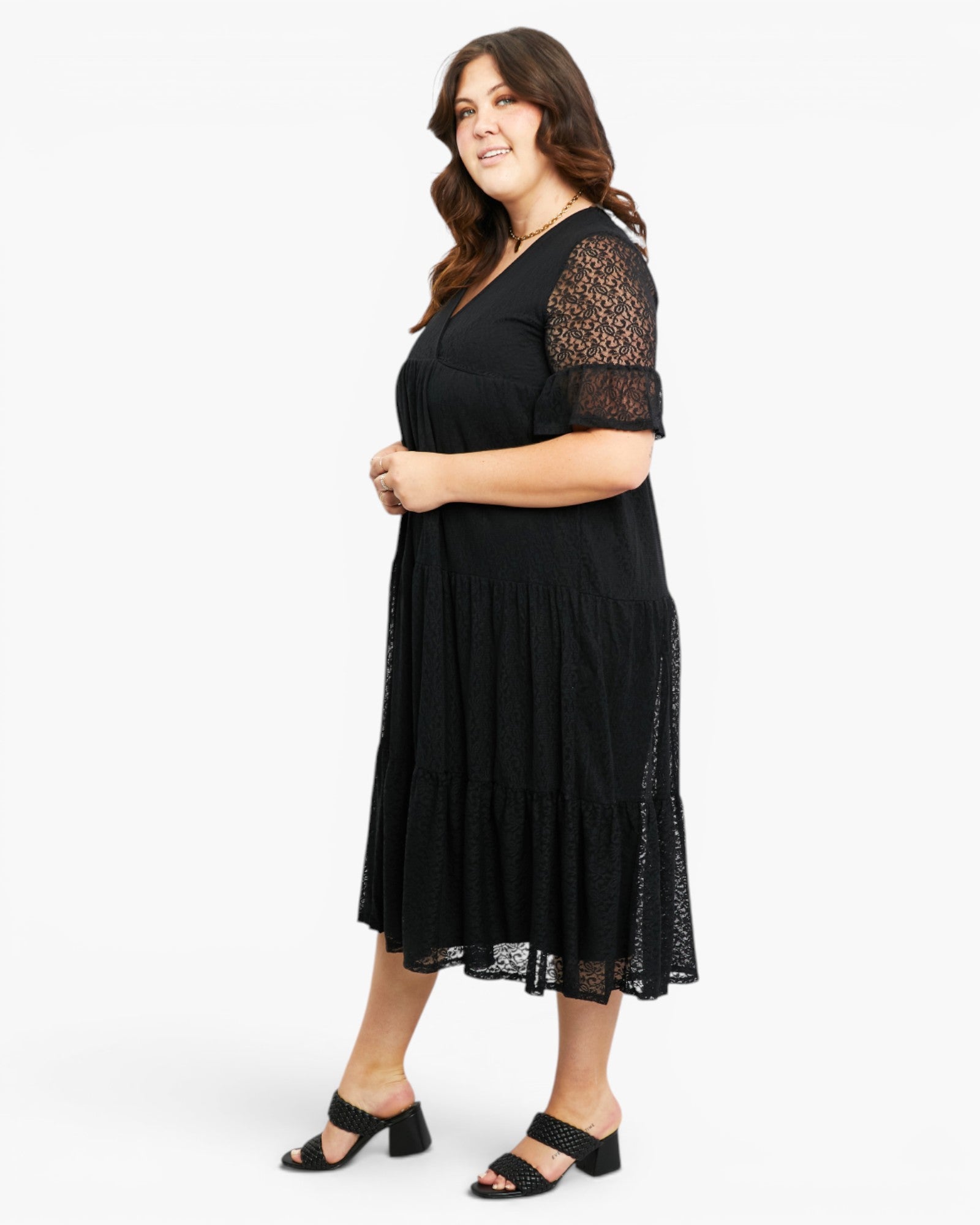 A woman modeling a black midi dress with sheer lace short sleeves and a tiered skirt, paired with black heeled sandals, against a plain white studio background.