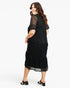 A woman modeling a black midi dress with sheer lace short sleeves and a tiered skirt, shown from a three-quarters back angle against a light gray studio background.