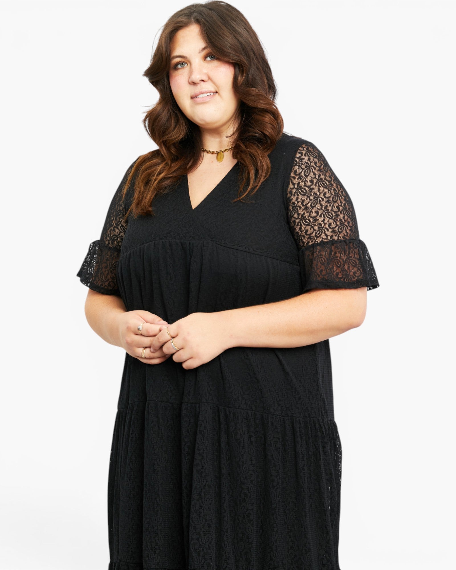 A woman modeling a black midi dress with short lace sleeves and a V-neckline, photographed against a plain light gray studio background.