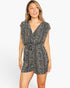 A woman modeling a short black and white patterned romper with a V-neck, cap sleeves, and a matching fabric belt with a square buckle, set against a plain light gray studio background.