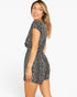 A woman modeling a short black romper with a white dalmatian-style spot pattern, shown from a three-quarters back angle against a plain white studio background.