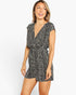 A woman modeling a short black and white patterned romper with a wrap-style V-neck and cap sleeves, photographed against a clean light gray studio background.