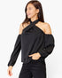 A woman wearing a black cold-shoulder long-sleeve blouse with a draped neckline and puffed sleeves, paired with black pants, against a light gray studio background.