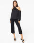 A woman modeling a black cold-shoulder top with long sleeves and black cropped pants, paired with black heeled sandals, against a plain white studio background.