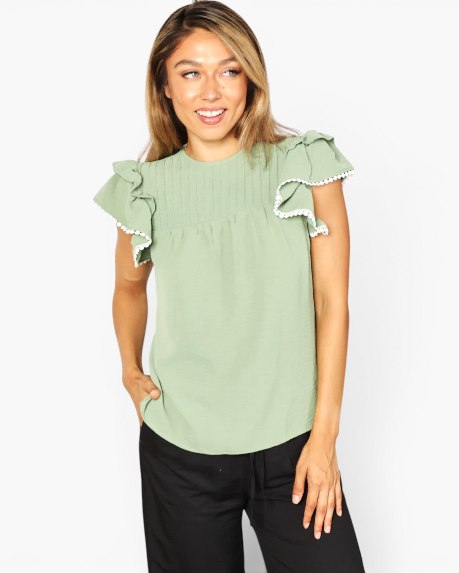 A woman wearing a sage green short-sleeve blouse with ruffled sleeves and white trim, paired with black pants, against a plain white background.