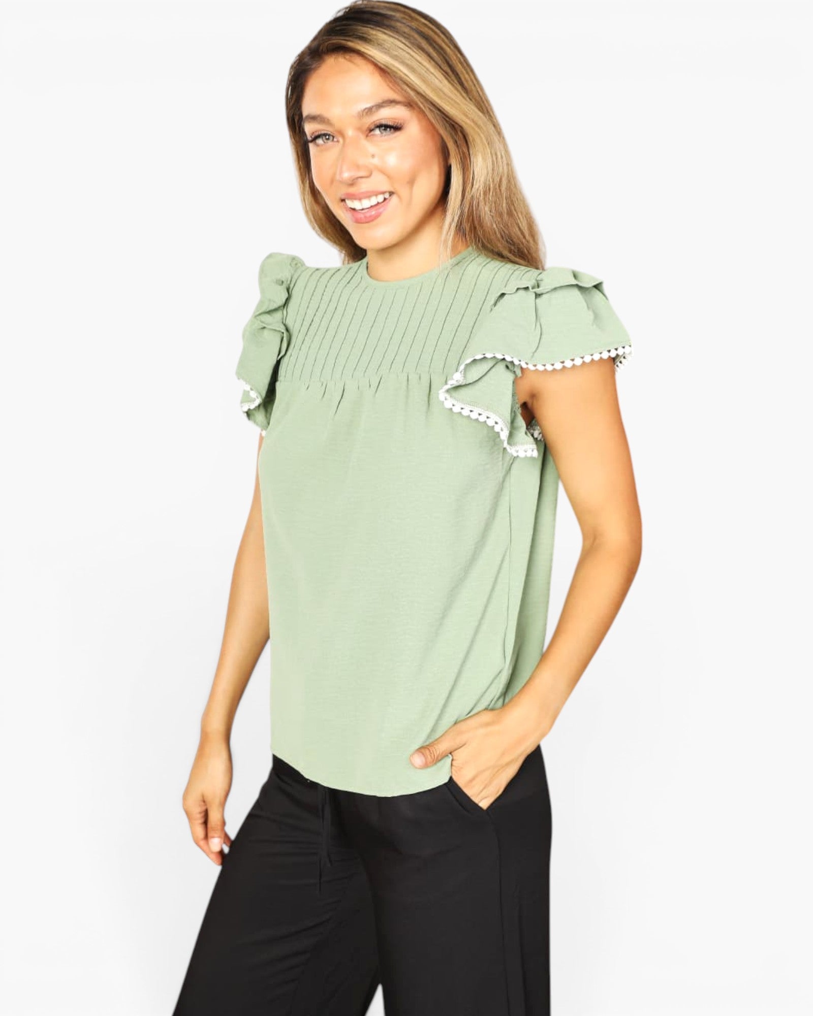 A woman wearing a sage green short-sleeve blouse with pleated detailing on the chest and ruffled sleeves finished with white pom-pom trim, paired with black pants, against a plain white background.