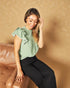 Woman wearing a green top with ruffled sleeves and black pants sitting on a brown couch against a beige wall.