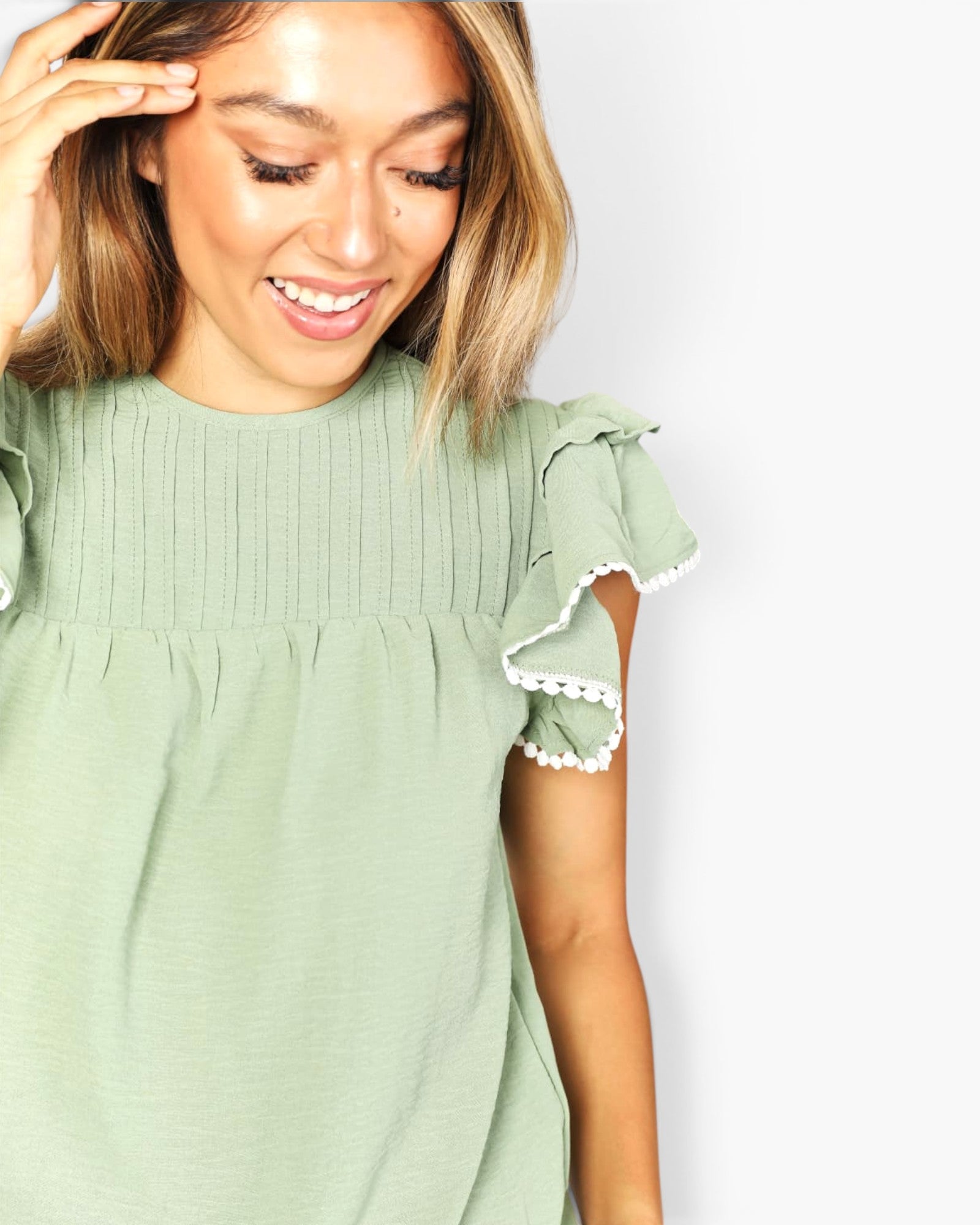 A woman wearing a sage green top with ruffled short sleeves and white pom-pom trim, shown against a plain light gray background.