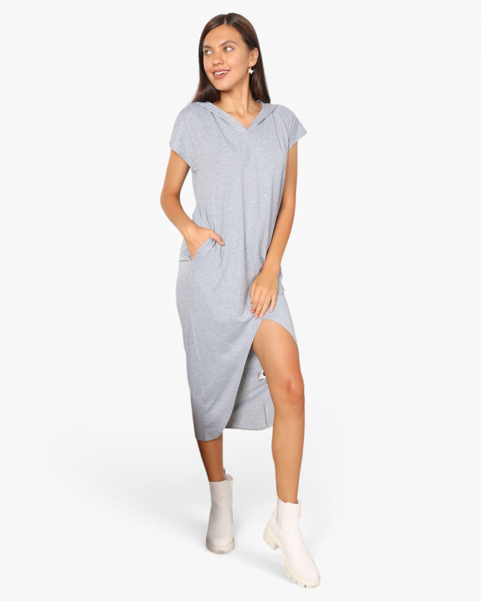 A woman modeling a light gray heathered short-sleeve hooded dress with a high side slit, paired with white ankle boots, against a plain white studio background.
