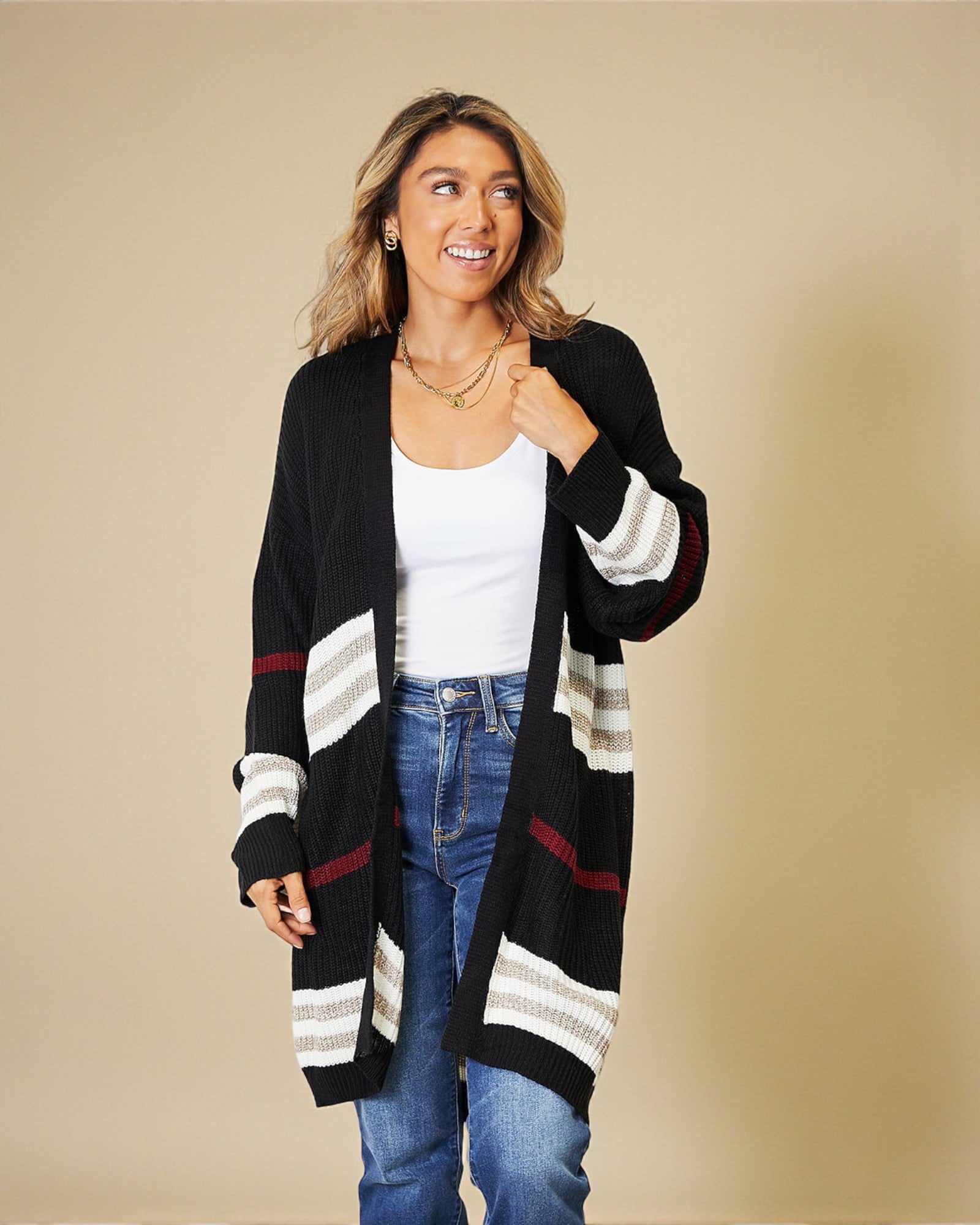 Woman wearing a black cardigan with white and red stripes, white top, and blue jeans on a beige background