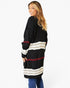 A woman modeling a black knit cardigan with horizontal stripes in cream, gray, and burgundy, paired with blue jeans against a plain white studio background.