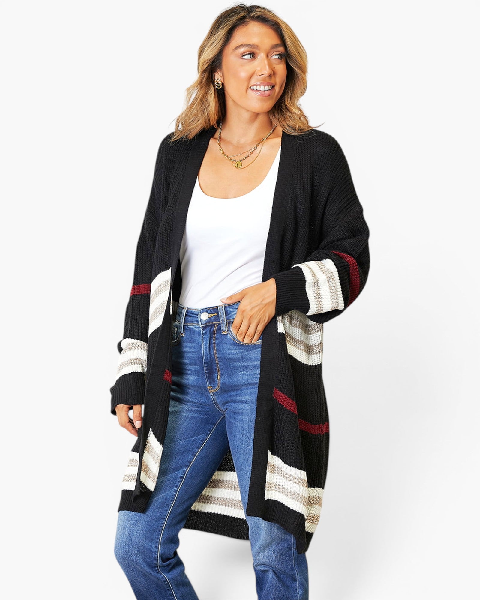 A woman modeling a long open-front knit cardigan with black, cream, and burgundy striped panels, worn over a white tank top and blue jeans against a light gray studio background.