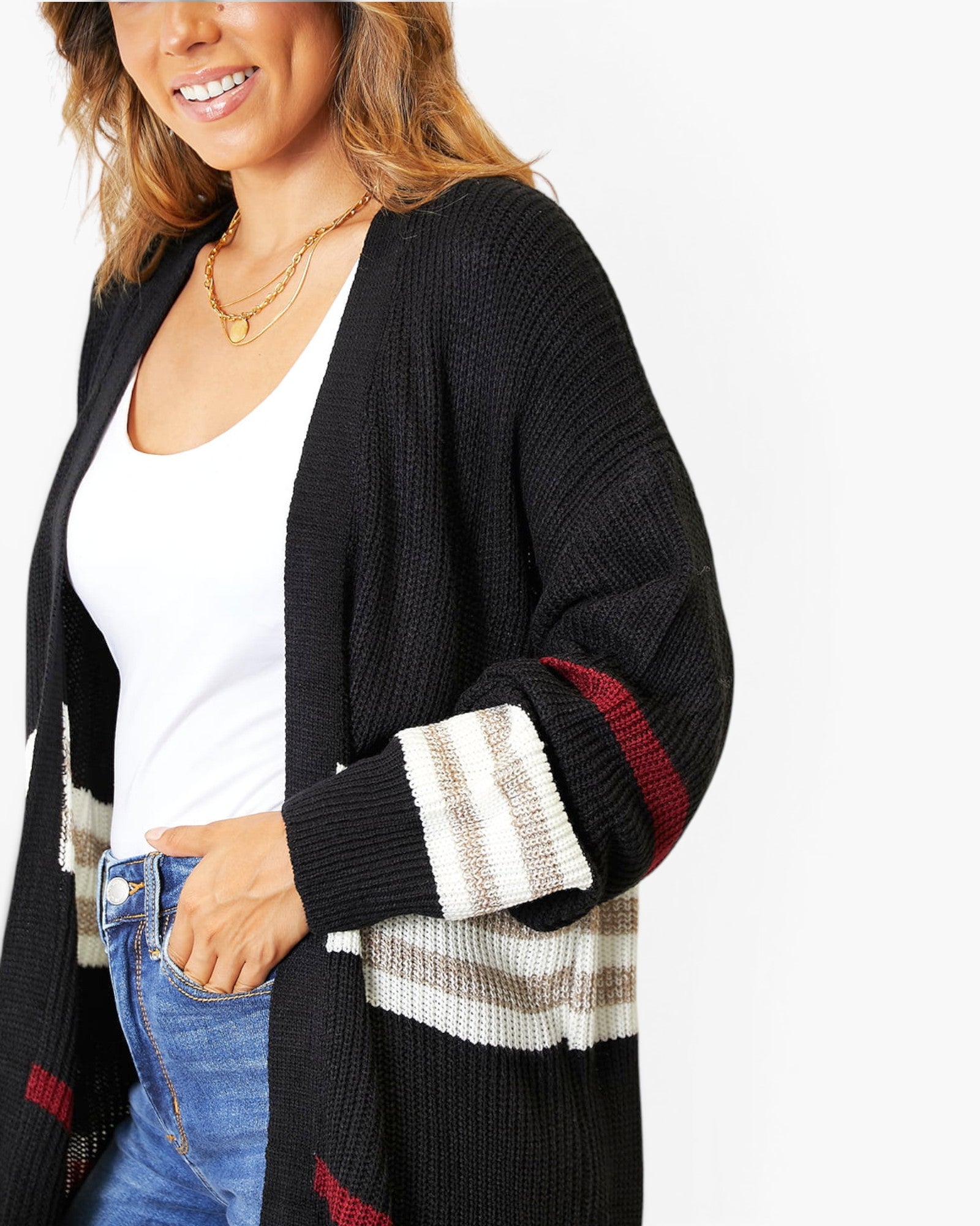 A woman wearing a black open-front cardigan with striped knit cuffs and a plaid pattern on the lower front, layered over a white tank top and blue jeans against a plain white background.