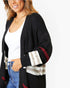 A woman wearing a black open-front cardigan with striped knit cuffs and a plaid pattern on the lower front, layered over a white tank top and blue jeans against a plain white background.