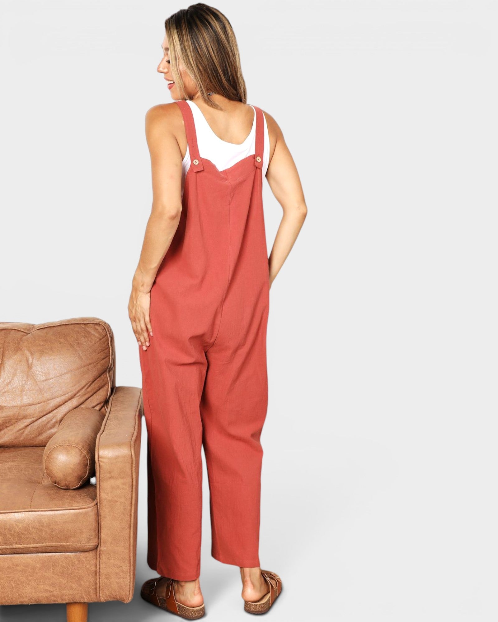 Woman wearing red overalls standing next to a brown leather chair on a plain background
