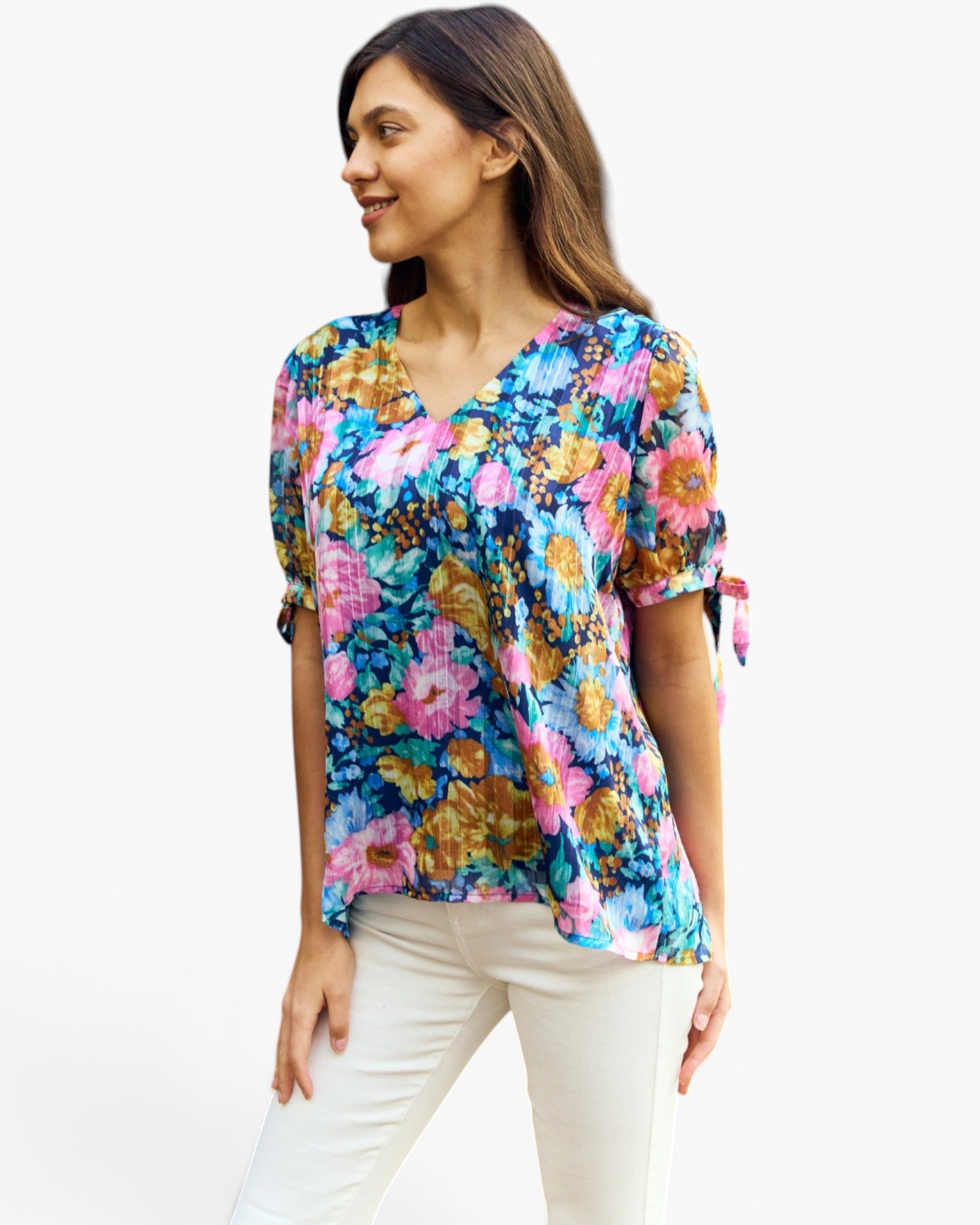 A woman wearing a vibrant, multicolor floral print blouse with short sleeves and tie details at the cuffs, paired with white pants, set against a plain white studio background.