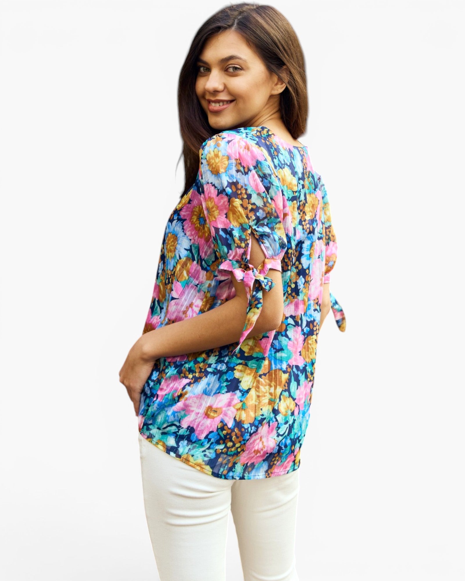 A woman wearing a colorful floral short-sleeve top with tie details on the sleeves and back, paired with white pants, photographed against a plain white background.