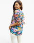 A woman wearing a colorful floral short-sleeve top with tie details on the sleeves and back, paired with white pants, photographed against a plain white background.