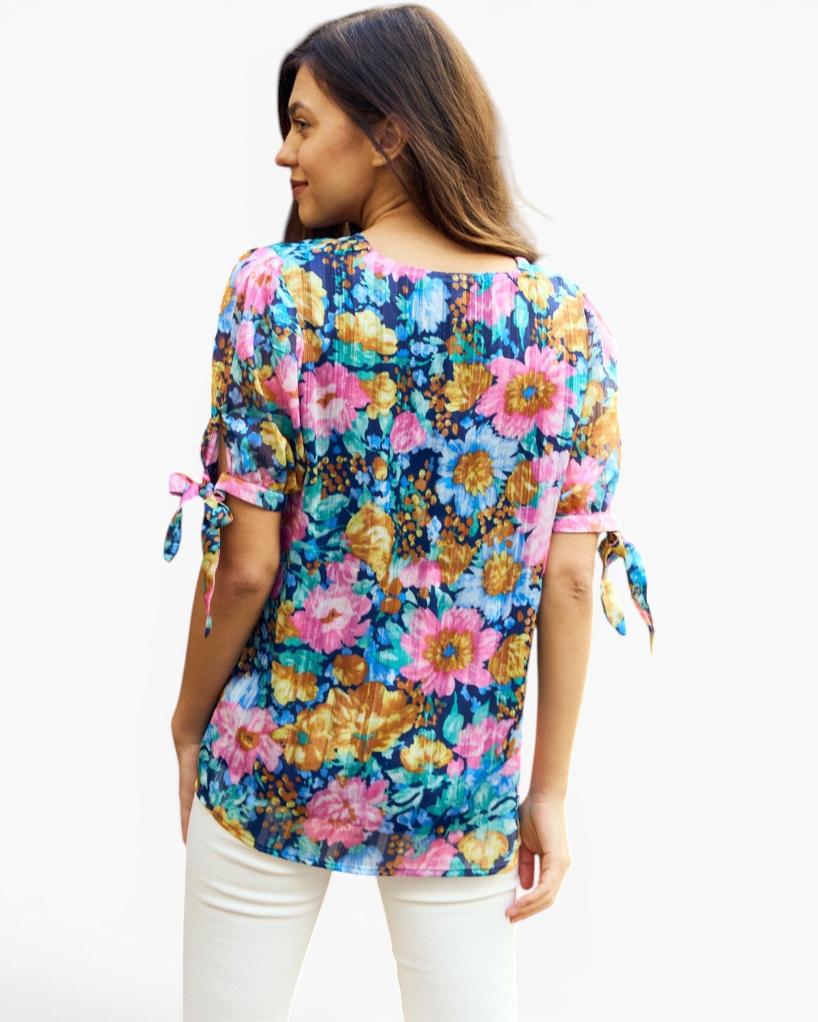 A woman shown from the back wearing a colorful floral print top with pink and blue tones, paired with white pants, against a plain white background.