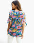 A woman shown from the back wearing a colorful floral print top with pink and blue tones, paired with white pants, against a plain white background.