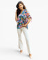 A woman modeling a colorful floral short-sleeve blouse with tie details at the cuffs, paired with white flared pants and beige heels, against a plain white studio background.